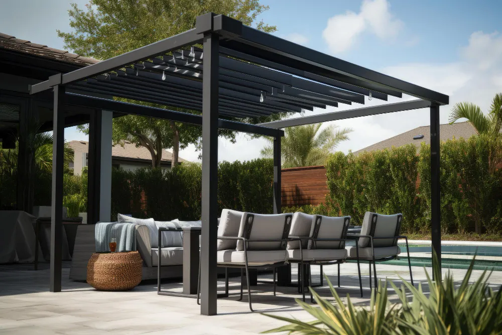 Modern outdoor patio with a black pergola, cushioned chairs, a wicker ottoman, and a pool with greenery in the background.